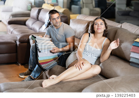 In store wife settled down with legs to test of sofa in use,husband examines upholstery fabric In store wife settled down with legs to test of sofa in use,husband examines upholstery fabric 139225403