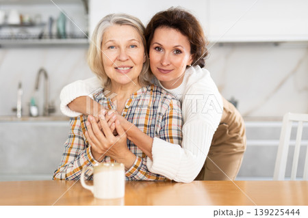 In kitchen, daughter and an mature mother hugging and drinking tea In kitchen, daughter and an mature mother hugging and drinking tea 139225444