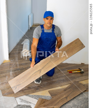Professional builder in blue construction overalls installing wooden laminate flooring, holding plank Professional builder in blue construction overalls installing wooden laminate flooring, holding plank 139225565