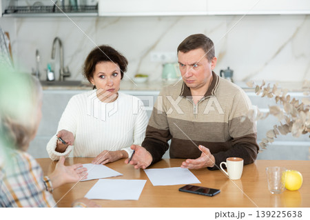 Middle-aged man and woman sitting at the kitchen-table face to face with female agent Middle-aged man and woman sitting at the kitchen-table face to face with female agent 139225638