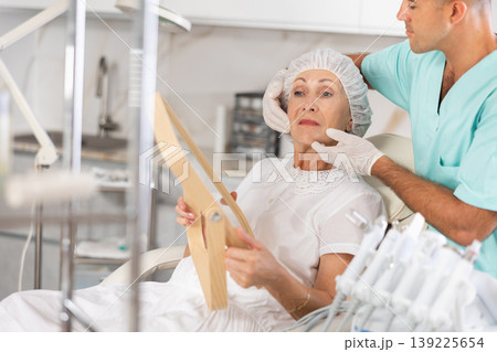 Male plastic surgeon giving professional advice after facial contouring to senior woman client looking at mirror Male plastic surgeon giving professional advice after facial contouring to senior woman client looking at mirror 139225654