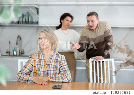 Upset old woman sitting at table with her back to angry middle-aged man and woman Upset old woman sitting at table with her back to angry middle-aged man and woman 139225681