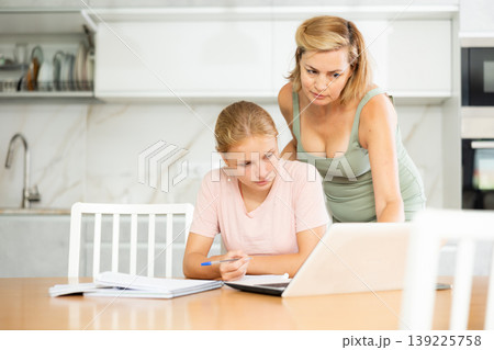 Mother looks at laptop and prompts and helps teen girl to write in notebook to complete task Mother looks at laptop and prompts and helps teen girl to write in notebook to complete task 139225758