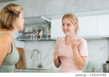 Cheerful teenage girl chatting friendly to mother in home kitchen Cheerful teenage girl chatting friendly to mother in home kitchen 139225766