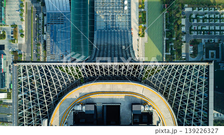 Aerial view of a modern building with a distinct patterned roof and curved facade, bordered by urban streets and a river during daytime Aerial view of a modern building with a distinct patterned roof and curved facade, bordered by urban streets and a river during daytime 139226327