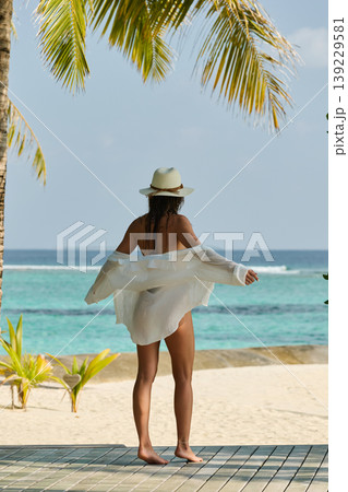 A beautiful tanned girl is wearing a hat and swimsuit with an athletic physique stands on the beach of the villa on a sunny day in the Maldives, she is happy and posing A beautiful tanned girl is wearing a hat and swimsuit with an athletic physique stands on the beach of the villa on a sunny day in the Maldives, she is happy and posing 139229581