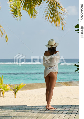 A beautiful tanned girl is wearing a hat and swimsuit with an athletic physique stands on the beach of the villa on a sunny day in the Maldives, she is happy and posing A beautiful tanned girl is wearing a hat and swimsuit with an athletic physique stands on the beach of the villa on a sunny day in the Maldives, she is happy and posing 139229596