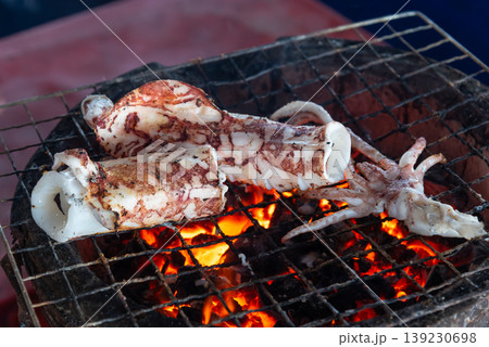 Close up of squid grilling over hot charcoal fire showing seafood cooking with smoky aroma crispy texture and street food style preparation Close up of squid grilling over hot charcoal fire showing seafood cooking with smoky aroma crispy texture and street food style preparation 139230698
