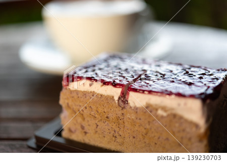Close up of berry cheesecake slice with cream layer and biscuit base topped with glossy fruit sauce served on table with blurred coffee cup in background Close up of berry cheesecake slice with cream layer and biscuit base topped with glossy fruit sauce served on table with blurred coffee cup in background 139230703