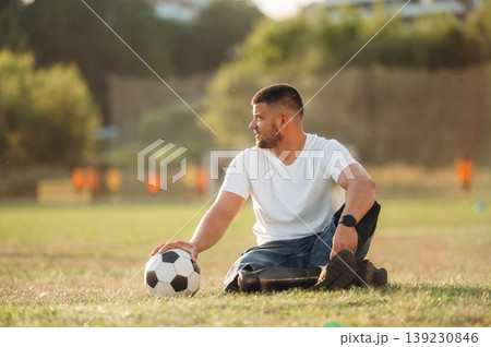 Motivation concept, sitting on the ground. Strong amputee man with prosthesis is with soccer ball on the field Motivation concept, sitting on the ground. Strong amputee man with prosthesis is with soccer ball on the field 139230846