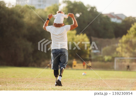 Outdoors, sitting on the shoulders. The amputee father is with his son on the field 139230954