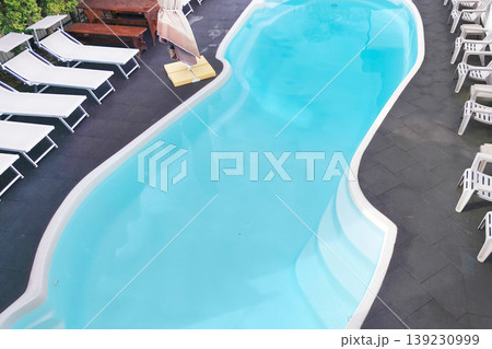 Top view of a curved turquoise swimming pool surrounded by loungers, chairs, and a shaded umbrella on a tiled patio, evoking a calm resort atmosphere and relaxation. Top view of a curved turquoise swimming pool surrounded by loungers, chairs, and a shaded umbrella on a tiled patio, evoking a calm resort atmosphere and relaxation. 139230999