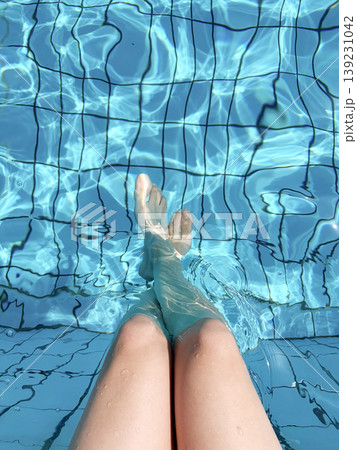 In the crystal clear, sunlit, sparkling blue water of the pool, the woman comfortably crossed her bare legs and feet. 139231042