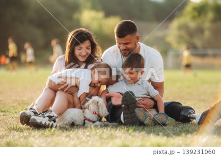 Resting by sitting on the ground. Mother, amputee father with prosthesis and kids are on the field Resting by sitting on the ground. Mother, amputee father with prosthesis and kids are on the field 139231060