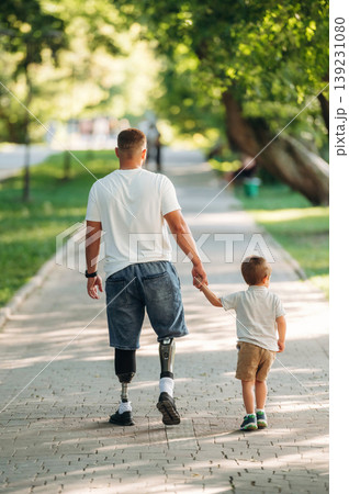 Rear view, from the back. Father that is amputee with prosthesis is with his little son in the park Rear view, from the back. Father that is amputee with prosthesis is with his little son in the park 139231080