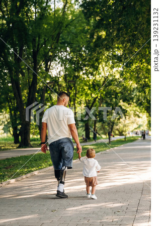 Rear view, from the back. Father that is amputee with prosthesis is with his little son in the park Rear view, from the back. Father that is amputee with prosthesis is with his little son in the park 139231132
