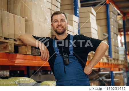 Leaning by the shelve, standing and posing. Man in uniform is working in the storage with packages for shipment Leaning by the shelve, standing and posing. Man in uniform is working in the storage with packages for shipment 139231432