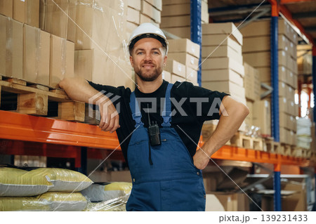 Leaning by the shelve, standing and posing. Man in uniform is working in the storage with packages for shipment Leaning by the shelve, standing and posing. Man in uniform is working in the storage with packages for shipment 139231433