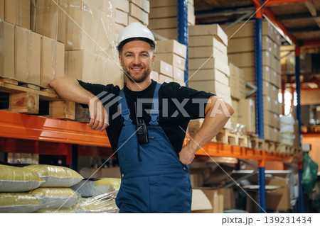 Leaning by the shelve, standing and posing. Man in uniform is working in the storage with packages for shipment Leaning by the shelve, standing and posing. Man in uniform is working in the storage with packages for shipment 139231434