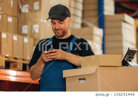 Texting by phone. Man in uniform is working in the storage with packages for shipment 139231444