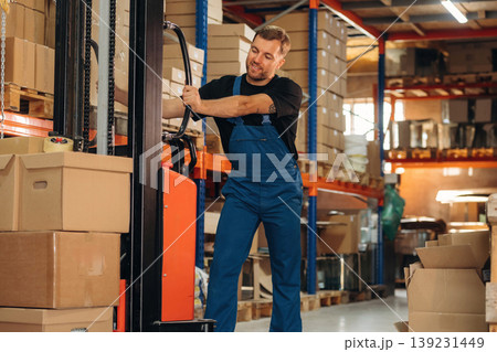 Moving stuff by using forklift. Man in uniform is working in the storage with packages for shipment Moving stuff by using forklift. Man in uniform is working in the storage with packages for shipment 139231449