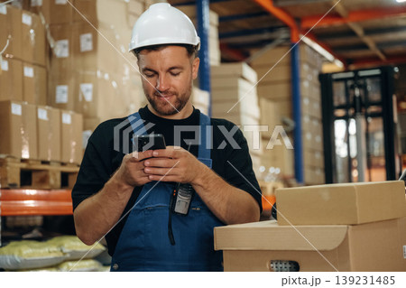 Uses smartphone, holding it in hands. Man in uniform is working in the storage with packages for shipment 139231485