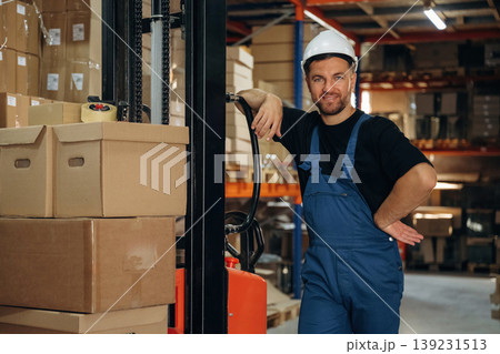 Model in hard hat is leaning on the forklift and smiling. Man in uniform is working in the storage with packages for shipment Model in hard hat is leaning on the forklift and smiling. Man in uniform is working in the storage with packages for shipment 139231513