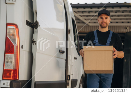 Looking forward, standing and holding the box. Delivery man is by the bus near garage outdoors Looking forward, standing and holding the box. Delivery man is by the bus near garage outdoors 139231524