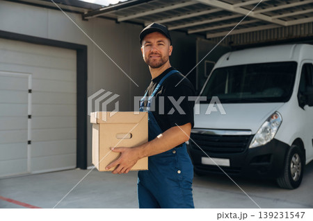 In black cap, with box in hands. Delivery man is by the bus near garage outdoors In black cap, with box in hands. Delivery man is by the bus near garage outdoors 139231547