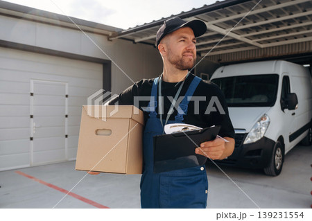 In black cap, with box in hands. Delivery man is by the bus near garage outdoors In black cap, with box in hands. Delivery man is by the bus near garage outdoors 139231554