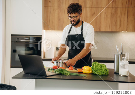 Laptop is on the table. Handsome man is preparing food on the kitchen Laptop is on the table. Handsome man is preparing food on the kitchen 139231808