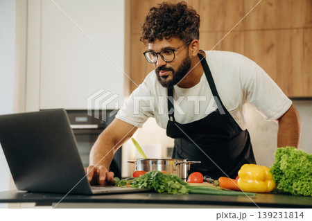 Taking online lessons, using laptop. Handsome man is preparing food on the kitchen Taking online lessons, using laptop. Handsome man is preparing food on the kitchen 139231814