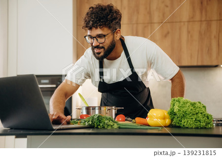 Taking online lessons, using laptop. Handsome man is preparing food on the kitchen Taking online lessons, using laptop. Handsome man is preparing food on the kitchen 139231815
