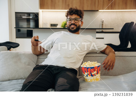 Popcorn and TV controller. Man is in the modern domestic room at home Popcorn and TV controller. Man is in the modern domestic room at home 139231839