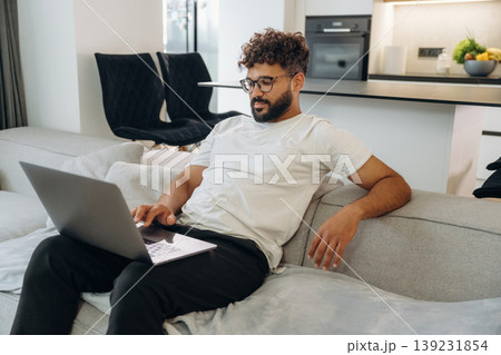 Laptop is on the knees, guy is sitting on the sofa. Man is in the modern domestic room at home Laptop is on the knees, guy is sitting on the sofa. Man is in the modern domestic room at home 139231854