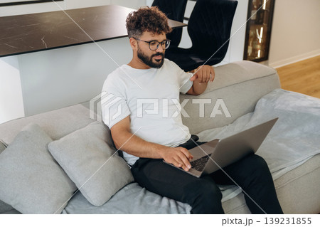 Laptop is on the knees, guy is sitting on the sofa. Man is in the modern domestic room at home Laptop is on the knees, guy is sitting on the sofa. Man is in the modern domestic room at home 139231855