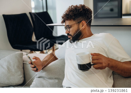 Cup of drink and smartphone are in hands. Man is in the modern domestic room at home Cup of drink and smartphone are in hands. Man is in the modern domestic room at home 139231861