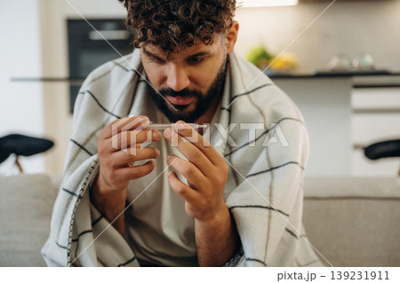 Thermometer is in hands, measuring the body temperature. Sick man is on the sofa at home, with towel, feeling bad Thermometer is in hands, measuring the body temperature. Sick man is on the sofa at home, with towel, feeling bad 139231911