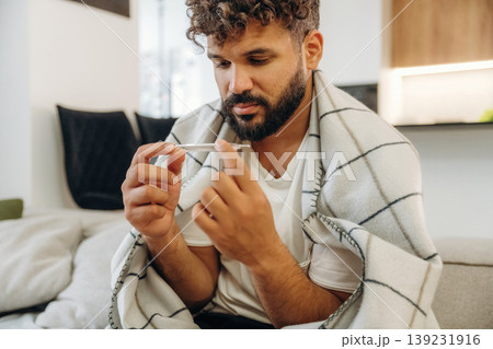 Body temperature measuring by using thermometer. Sick man is on the sofa at home, with towel, feeling bad 139231916