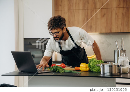 Laptop is on the table. Handsome man is preparing food on the kitchen Laptop is on the table. Handsome man is preparing food on the kitchen 139231996