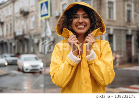 In yellow raincoat, standing and smiling. Happy young woman in the city during the rain In yellow raincoat, standing and smiling. Happy young woman in the city during the rain 139232175