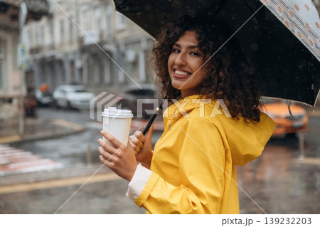 Hot drink is in the paper cup. Happy young woman in the city during the rain Hot drink is in the paper cup. Happy young woman in the city during the rain 139232203