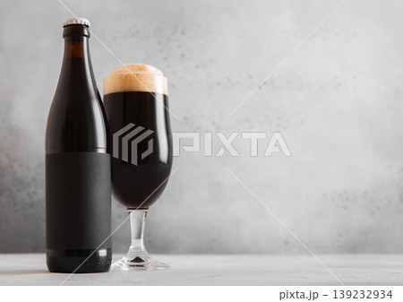 Bottle and glass of fresh cold dark stout beer with foam on light table background. Bottle and glass of fresh cold dark stout beer with foam on light table background. 139232934