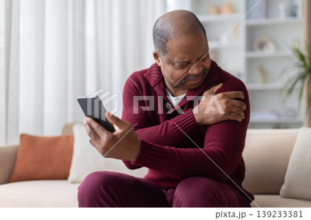 Mature black man grimacing in shoulder pain while holding smartphone at home 139233381