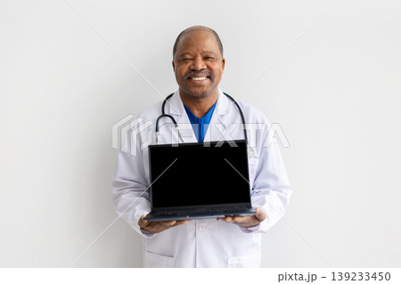 Smiling mature black doctor in white coat holding laptop with blank screen 139233450
