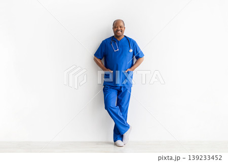 Confident mature black male doctor in blue scrubs leaning against white wall 139233452