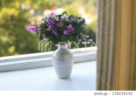 Bouquet of purple flowers in a small iridescent vase on a windowsill 139234242