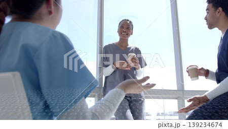 Chatting medical team in scrubs holding two paper coffee cups by floor-to-ceiling windows in lounge Chatting medical team in scrubs holding two paper coffee cups by floor-to-ceiling windows in lounge 139234674