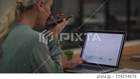 Holding smartphone near mouth, woman analyzing laptop chart at office desk with printed charts Holding smartphone near mouth, woman analyzing laptop chart at office desk with printed charts 139234678