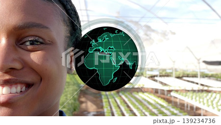 Smiling woman showing hydroponic trays in greenhouse, with green holographic globe, copy space 139234736